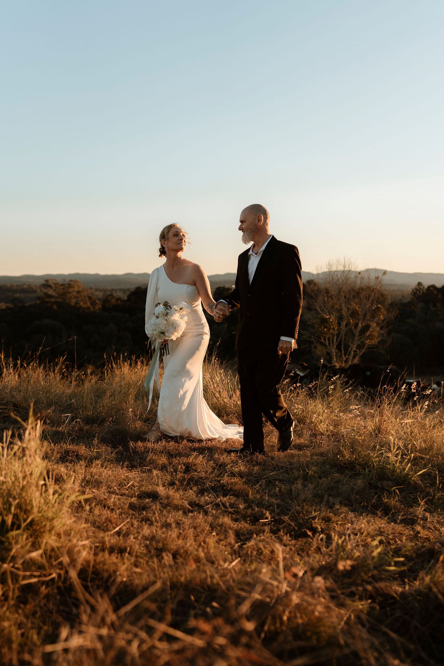 Bride and groom hold hands at golden hour on a grassy hilltop, with sweeping landscape views behind them.
