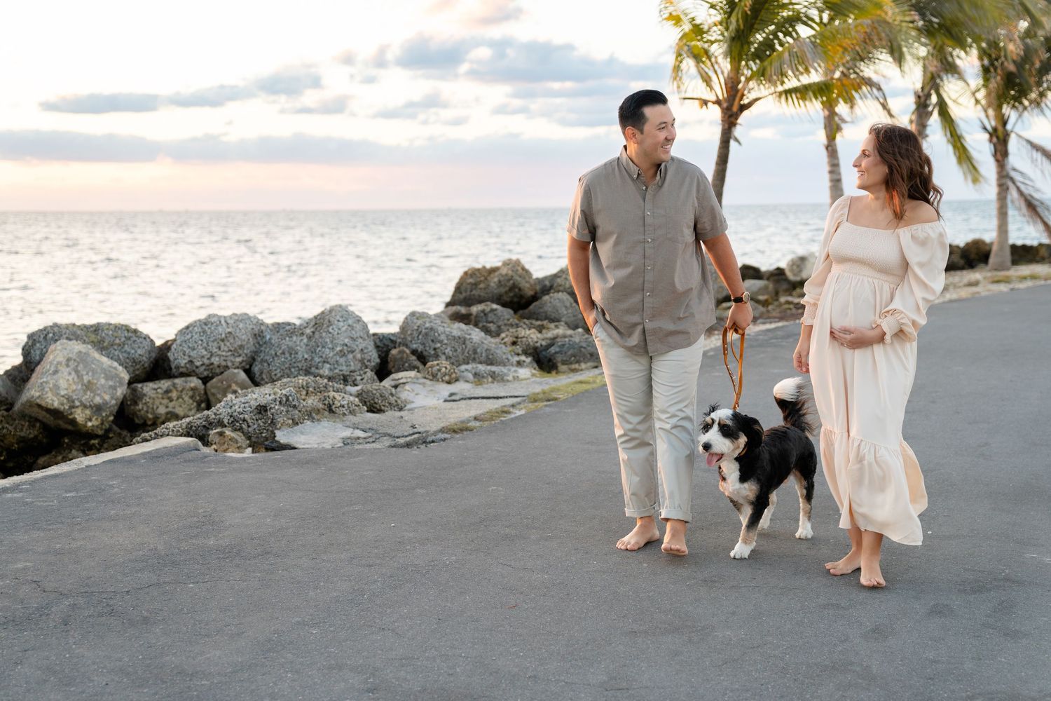 Pregnant woman in white dress walks with partner and border collie dog along coastal path with palm trees at sunset.