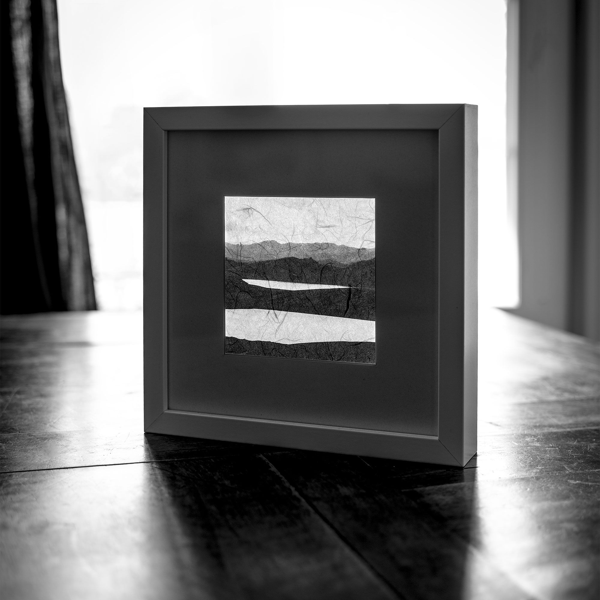 Black and white framed art print of a minimalist mountain landscape, displayed on a dark wooden surface.