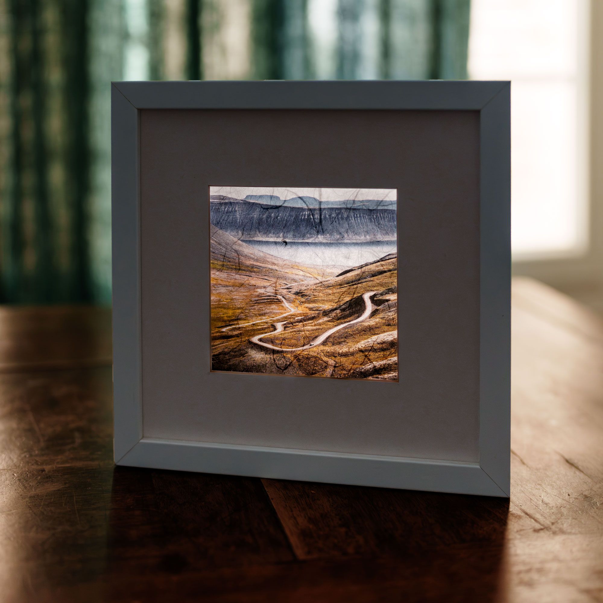 Framed fine art print of a winding mountain road through a vast valley, displayed on a wooden surface.