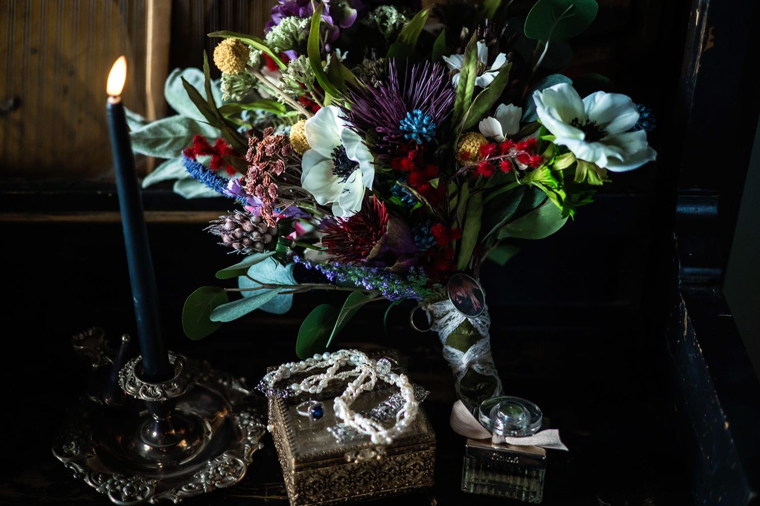 Dark floral wedding bouquet with anemones and berries beside a candle and jewelry box.