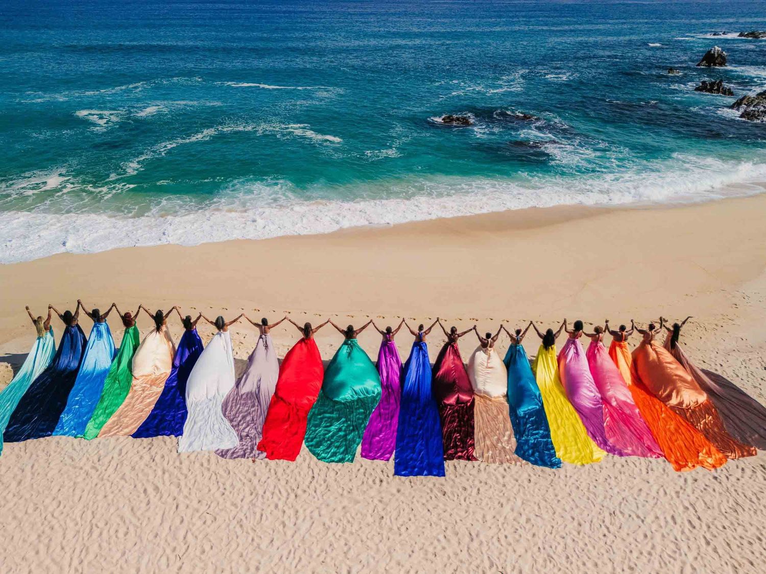 Colorful rainbow dresses displayed on a sandy beach with turquoise ocean waves in the background.
