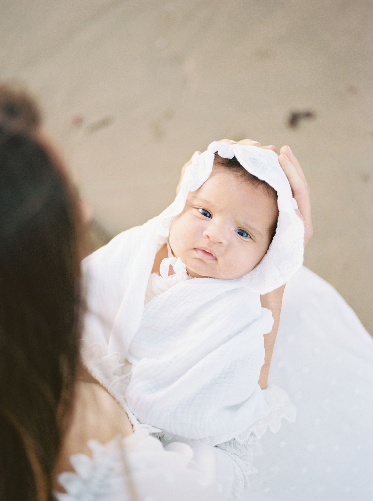 A newborn baby wrapped in a white swaddle and bonnet being held by a mother on a sandy beach.