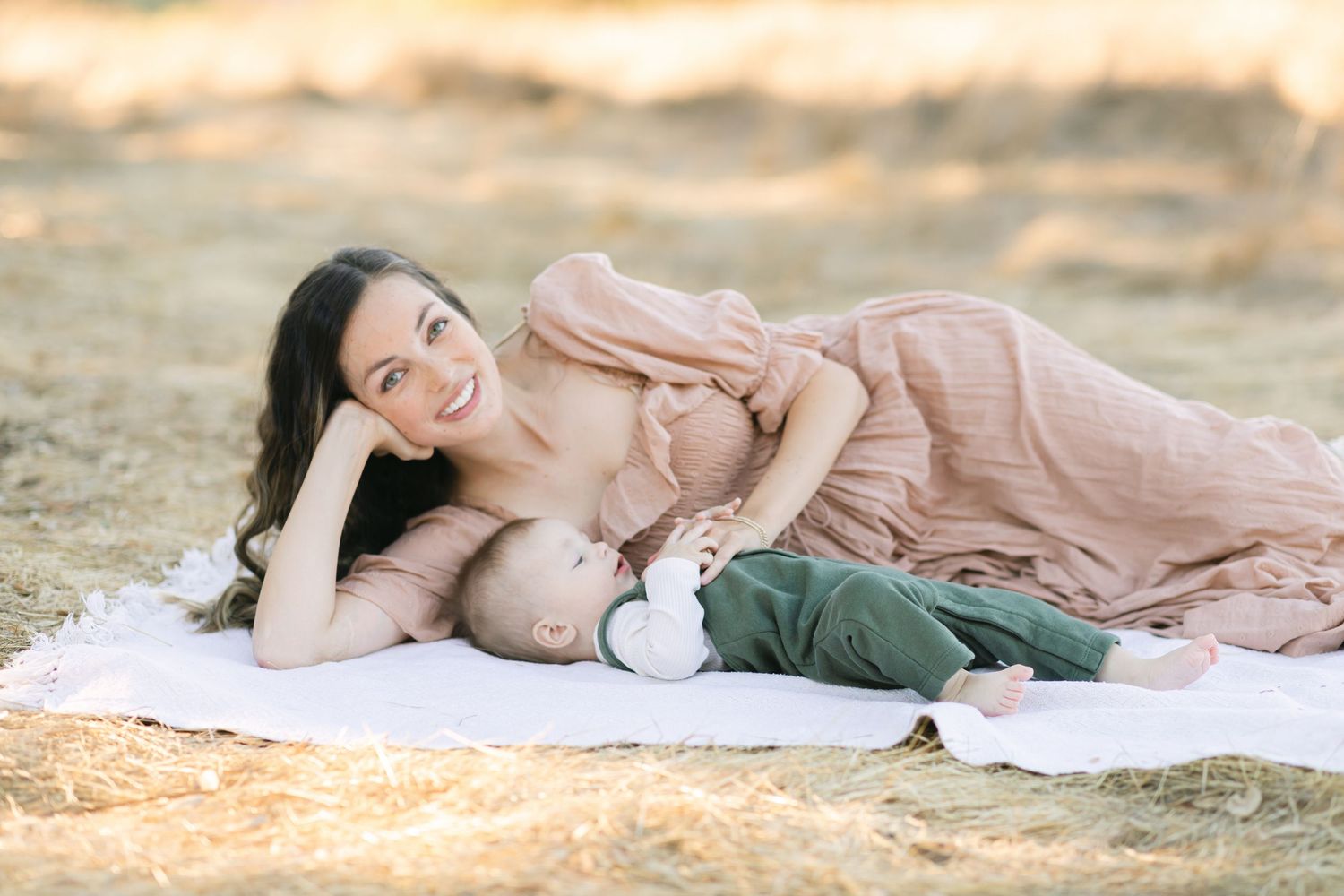 Mother in pink dress lying on blanket outdoors, breastfeeding her infant in green pants in a golden field.