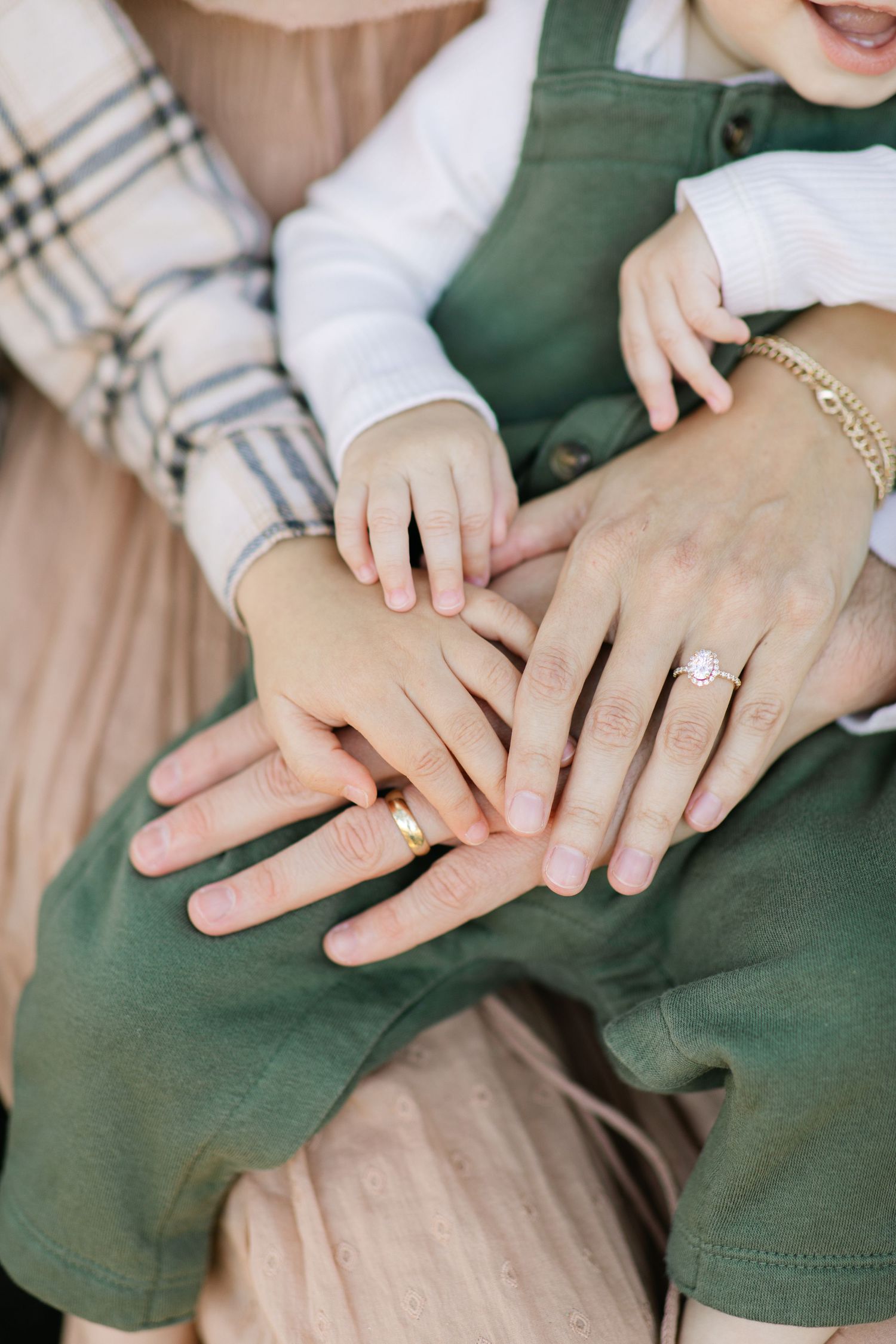 Family hands stacked together showing wedding rings and bracelet, with a baby in green overalls.