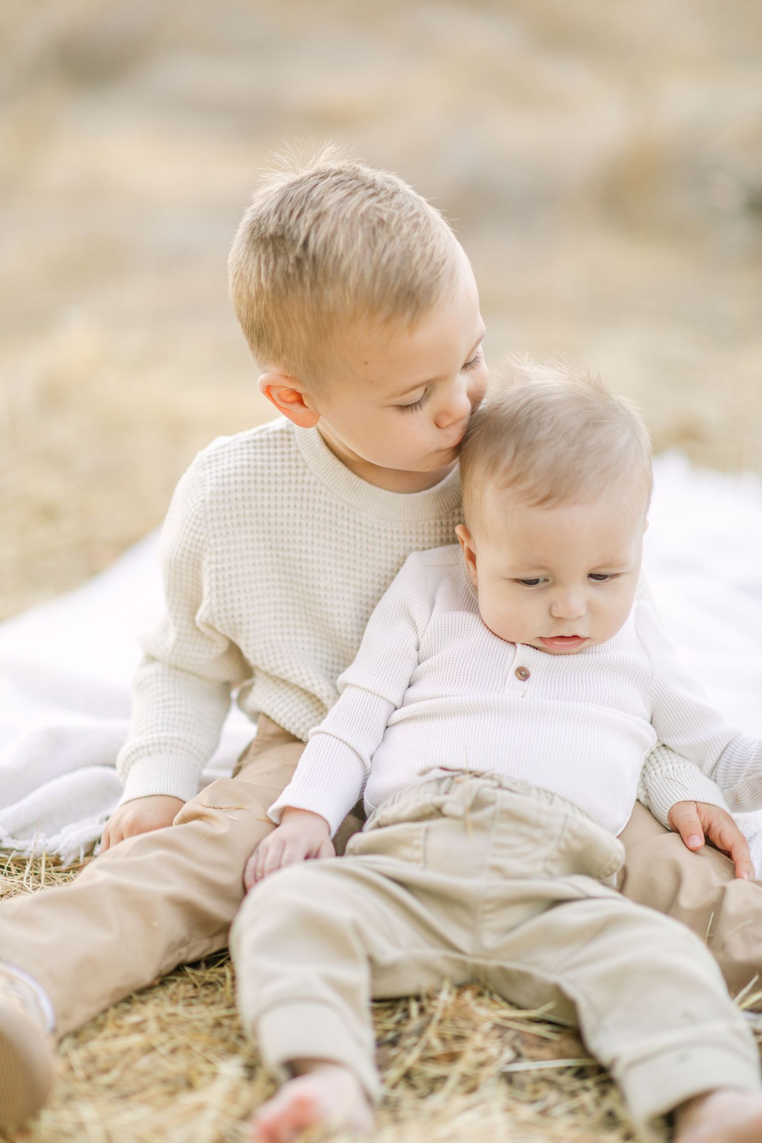 Older toddler boy kissing baby sibling on head while sitting together on white blanket in hay field.