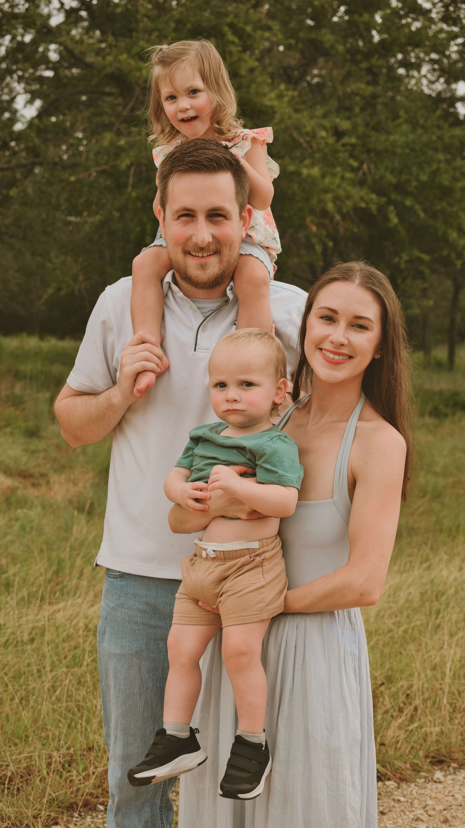 A smiling family of four poses outdoors, with a man holding a girl on his shoulders and a woman carrying a toddler.