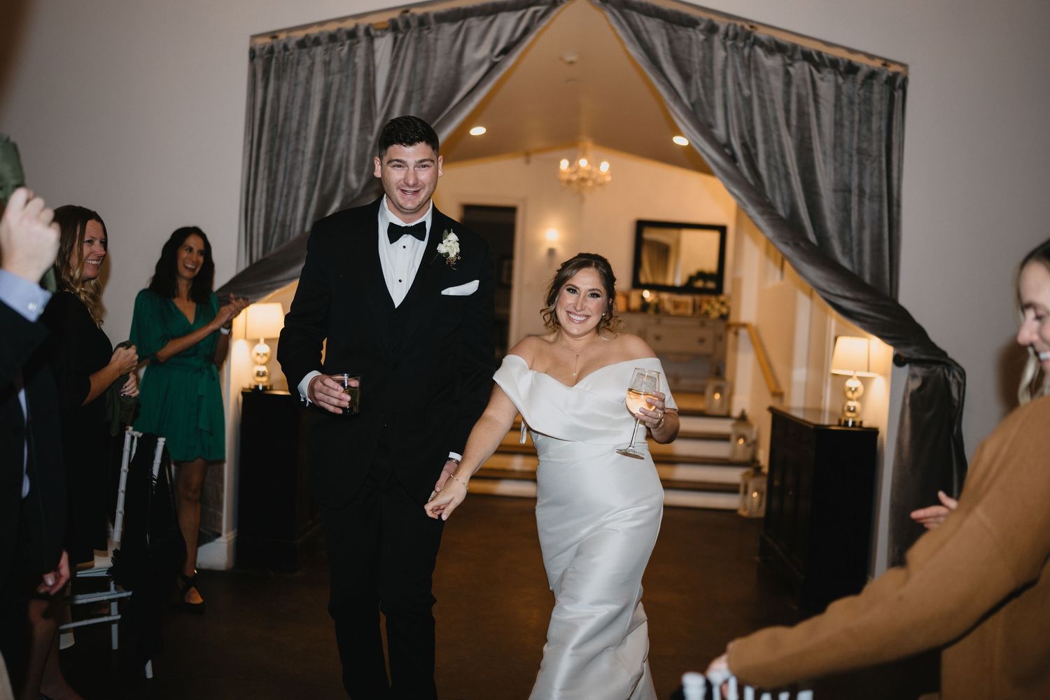 Bride in off-shoulder gown and groom in black tuxedo make grand entrance at elegant wedding reception.