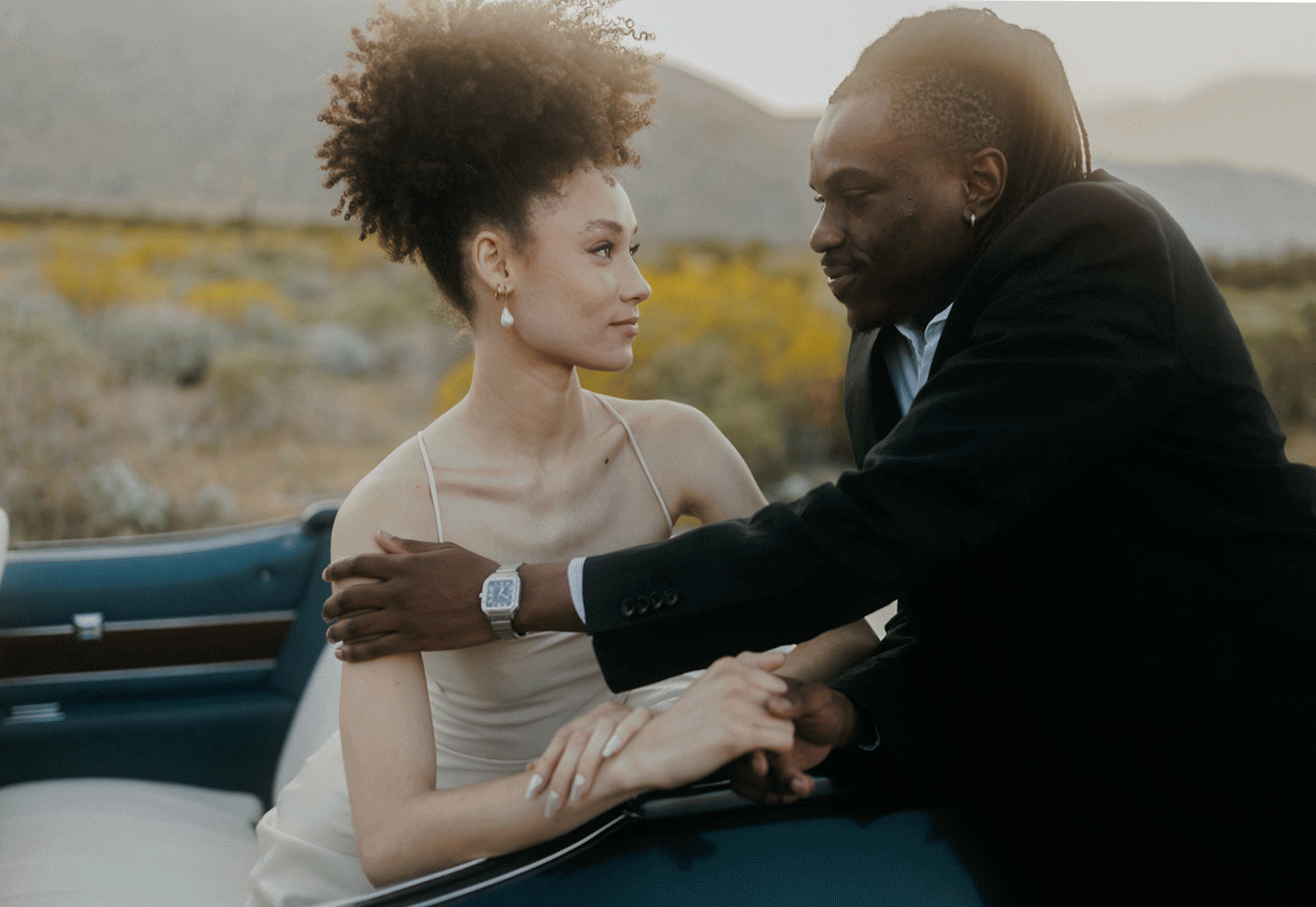 A couple shares an intimate moment while sitting in a convertible car in a desert setting at dusk.