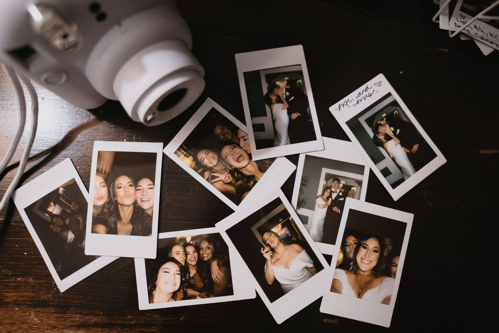 Polaroid photos of smiling women scattered on a dark wooden table next to a white Instax mini camera.