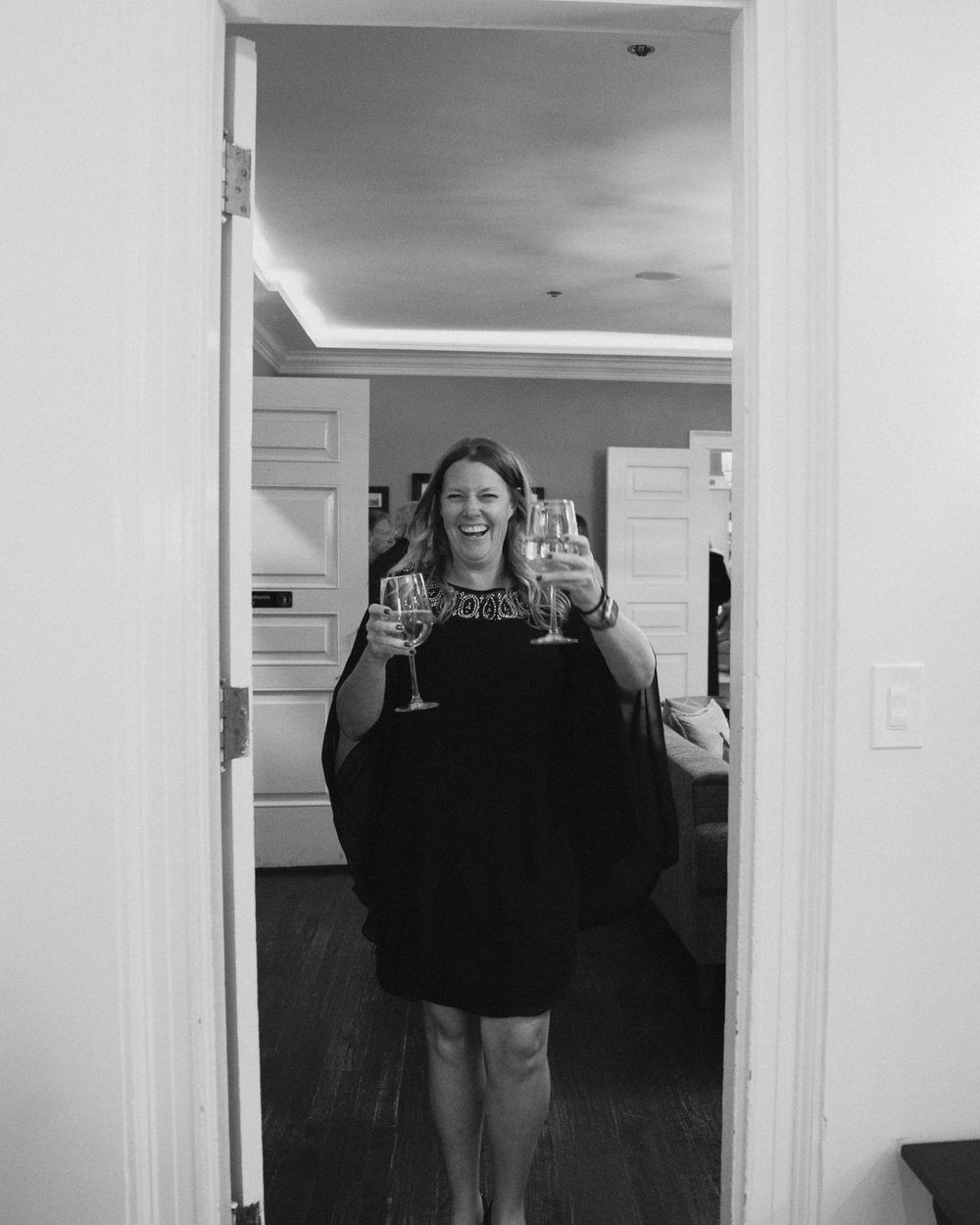 Woman in black dress laughing and holding wine glasses in a doorway, black and white photo.