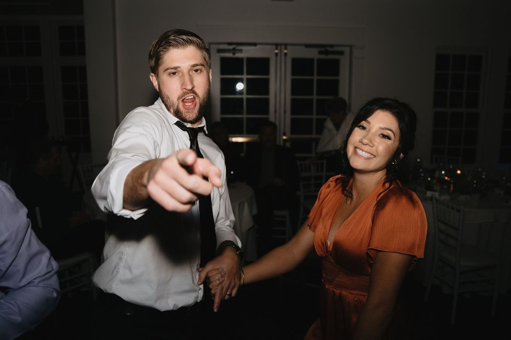 Man in white shirt pointing at camera while woman in orange dress smiles at wedding reception dance floor.