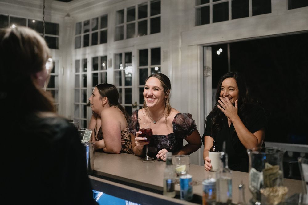 Women laugh and chat at a bar during an evening wedding reception.