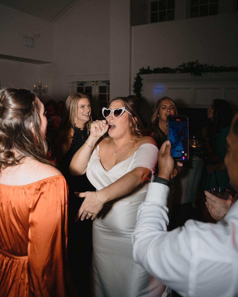 Bride in white dress and heart sunglasses singing at wedding reception while guests dance and celebrate around her.