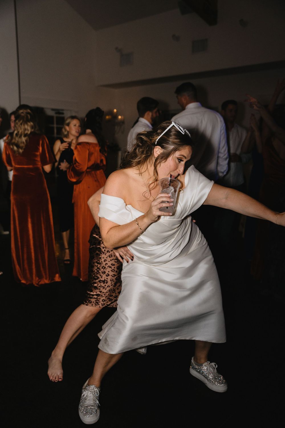 Bride dances energetically in white dress and sneakers while holding a drink at wedding reception.