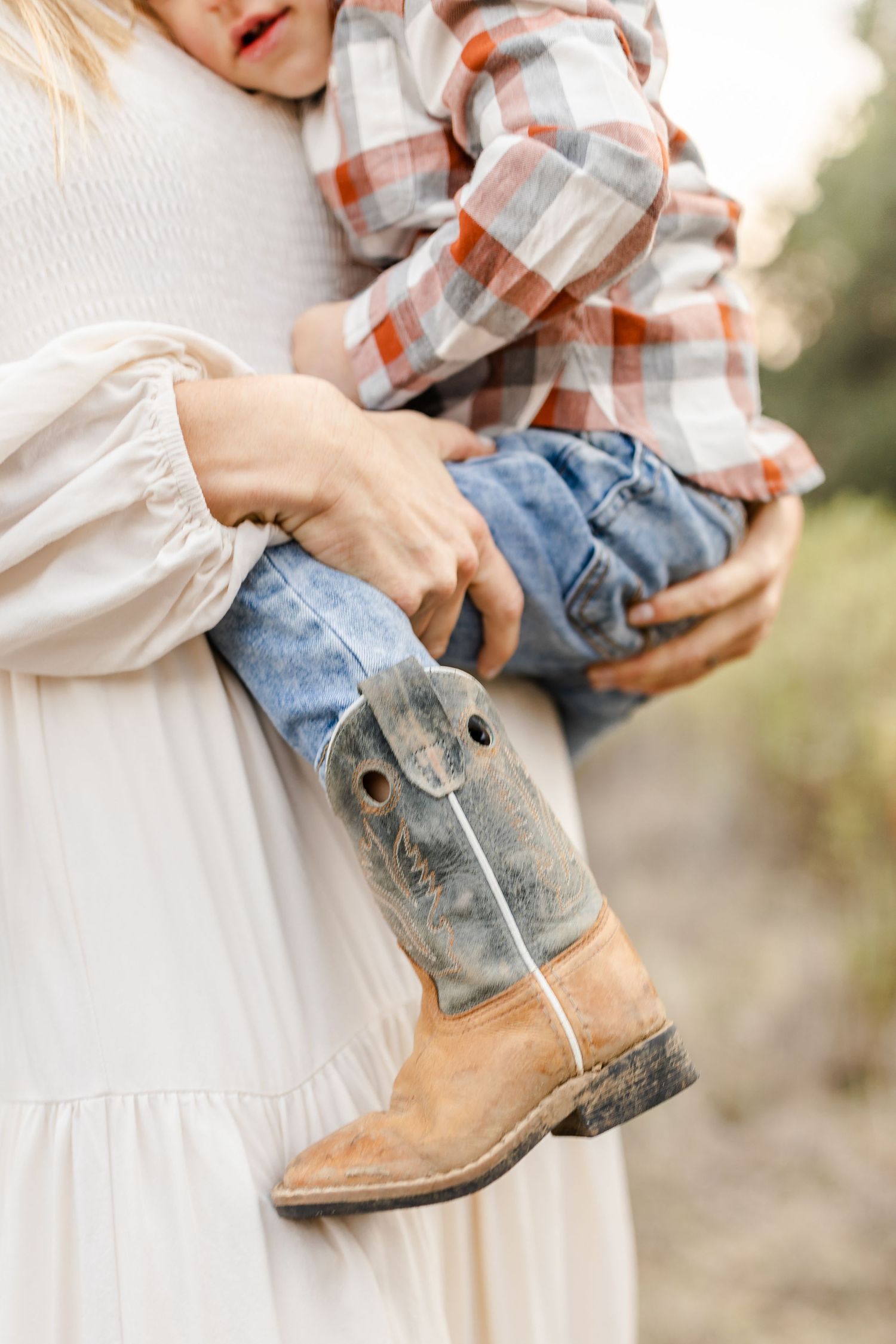 A mother in a white dress holds a toddler wearing a plaid shirt, jeans, and worn cowboy boots outdoors.