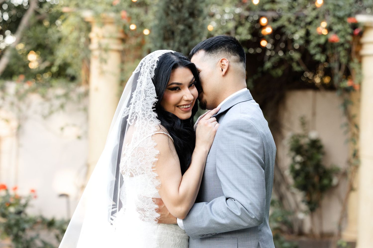 Bride in lace veil and groom in gray suit share romantic moment outdoors at elegant wedding venue.