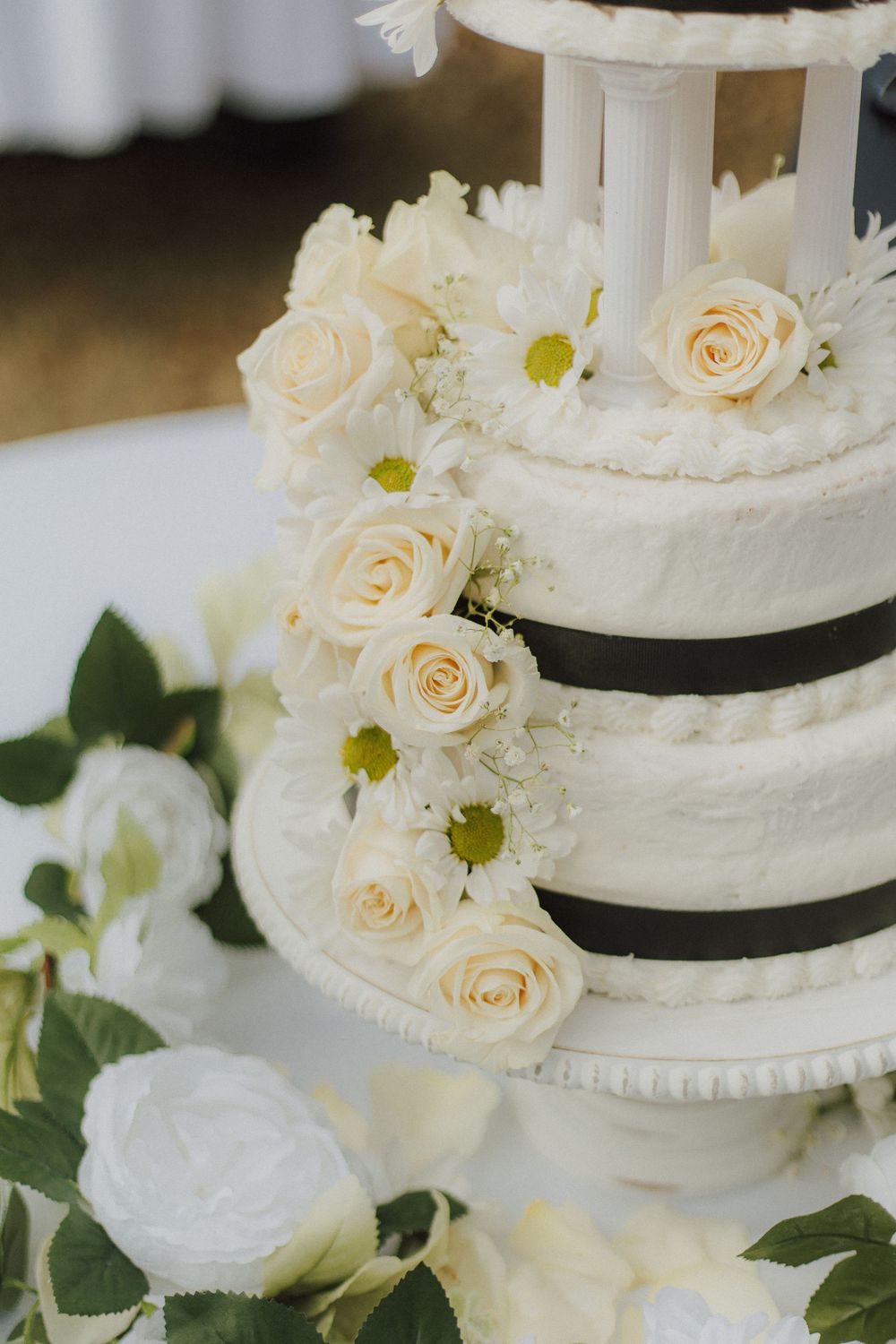 Elegant white wedding cake decorated with cream roses and daisies, featuring black ribbon accents between tiers.