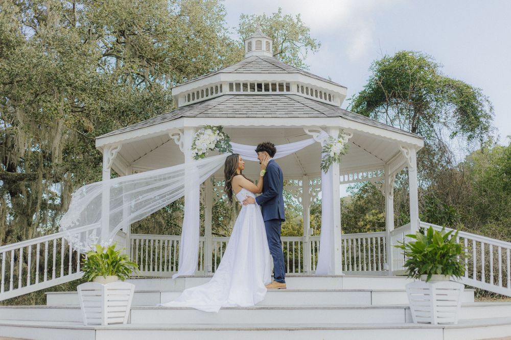A white wedding gazebo surrounded by lush trees and landscaping in a romantic outdoor setting.