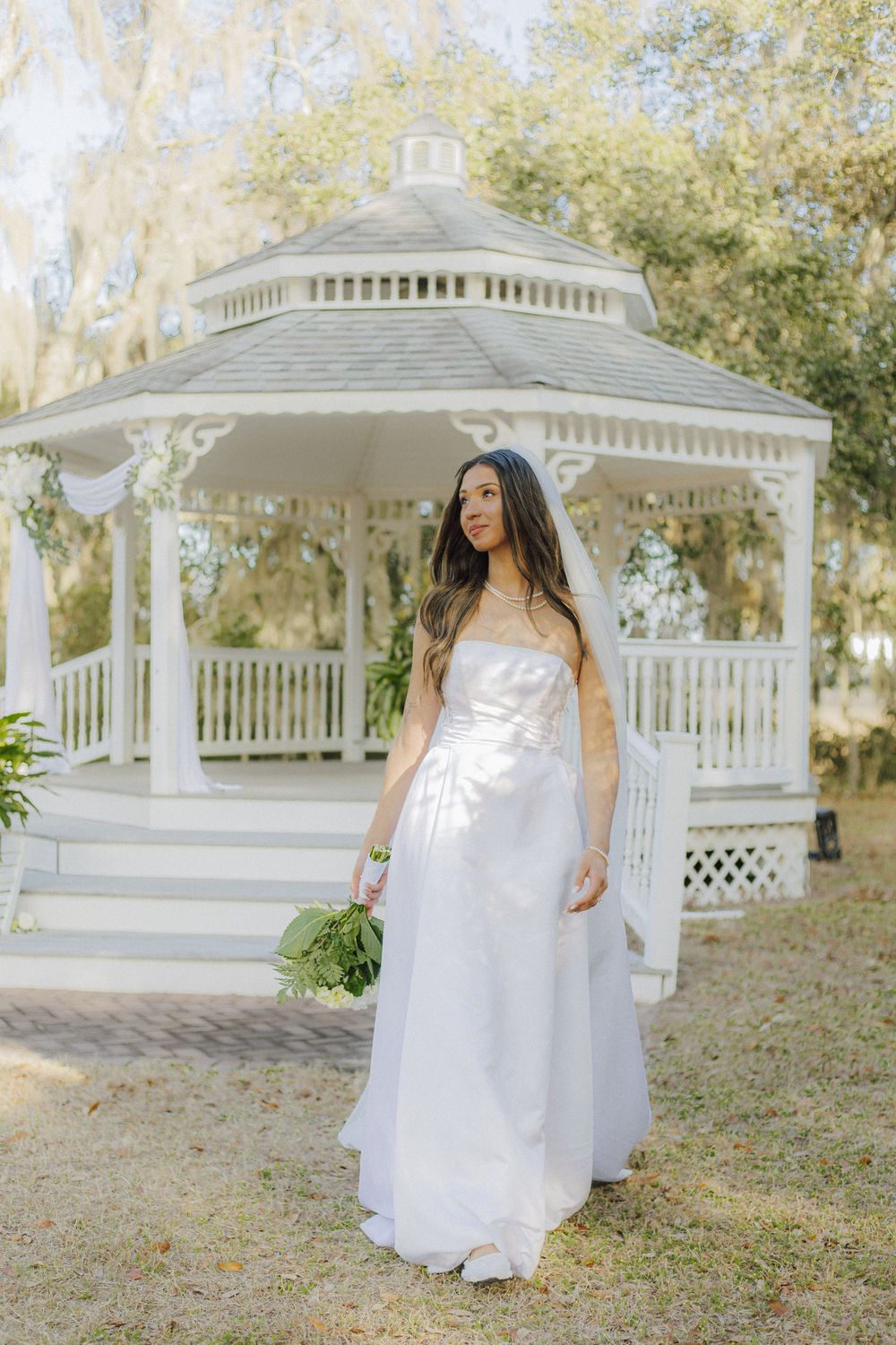 A white gazebo in a garden setting provides a romantic backdrop for a bride in a flowing white wedding dress.