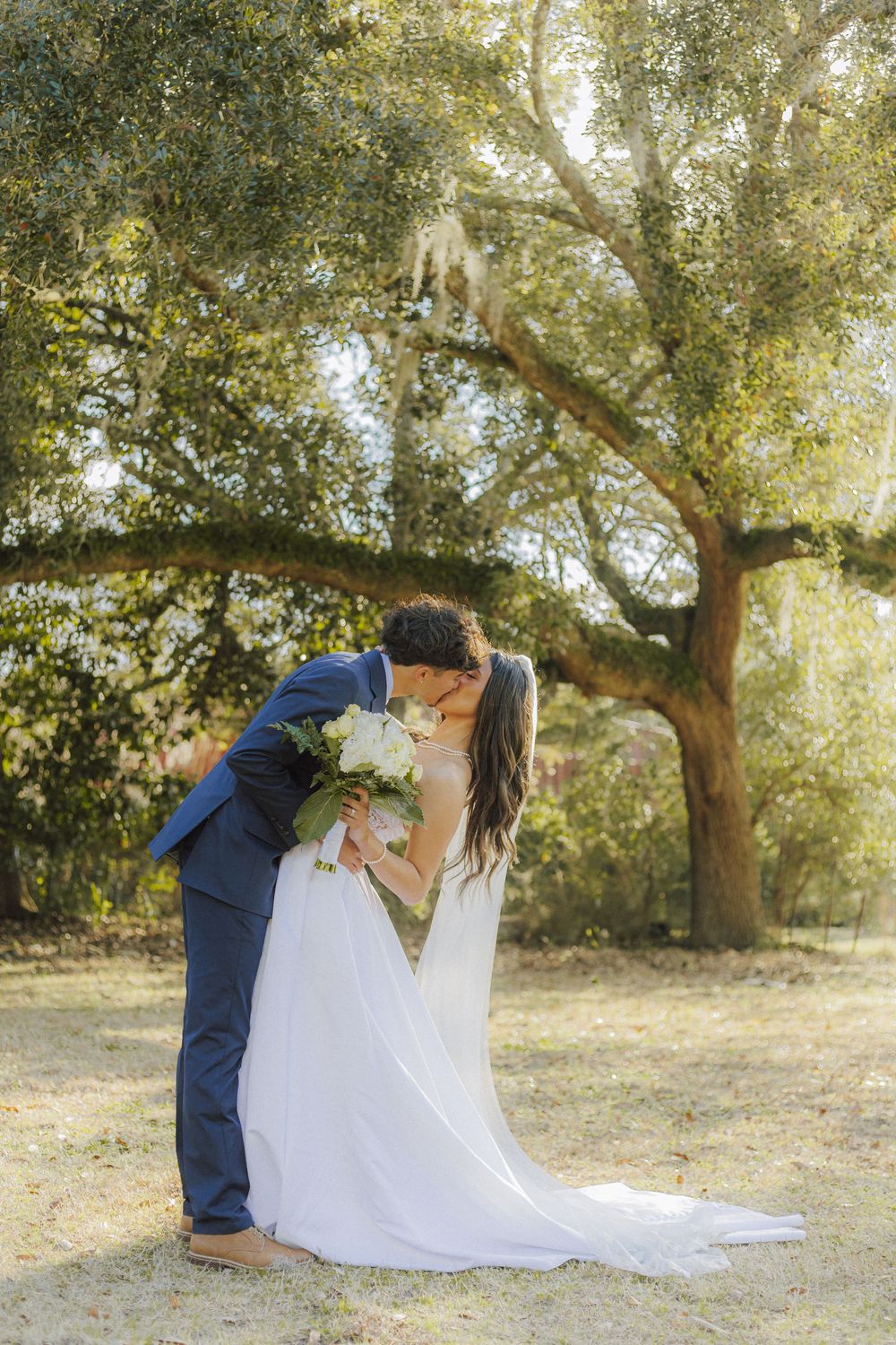Romantic outdoor wedding photo shoot with couple embracing under sunlit oak trees.