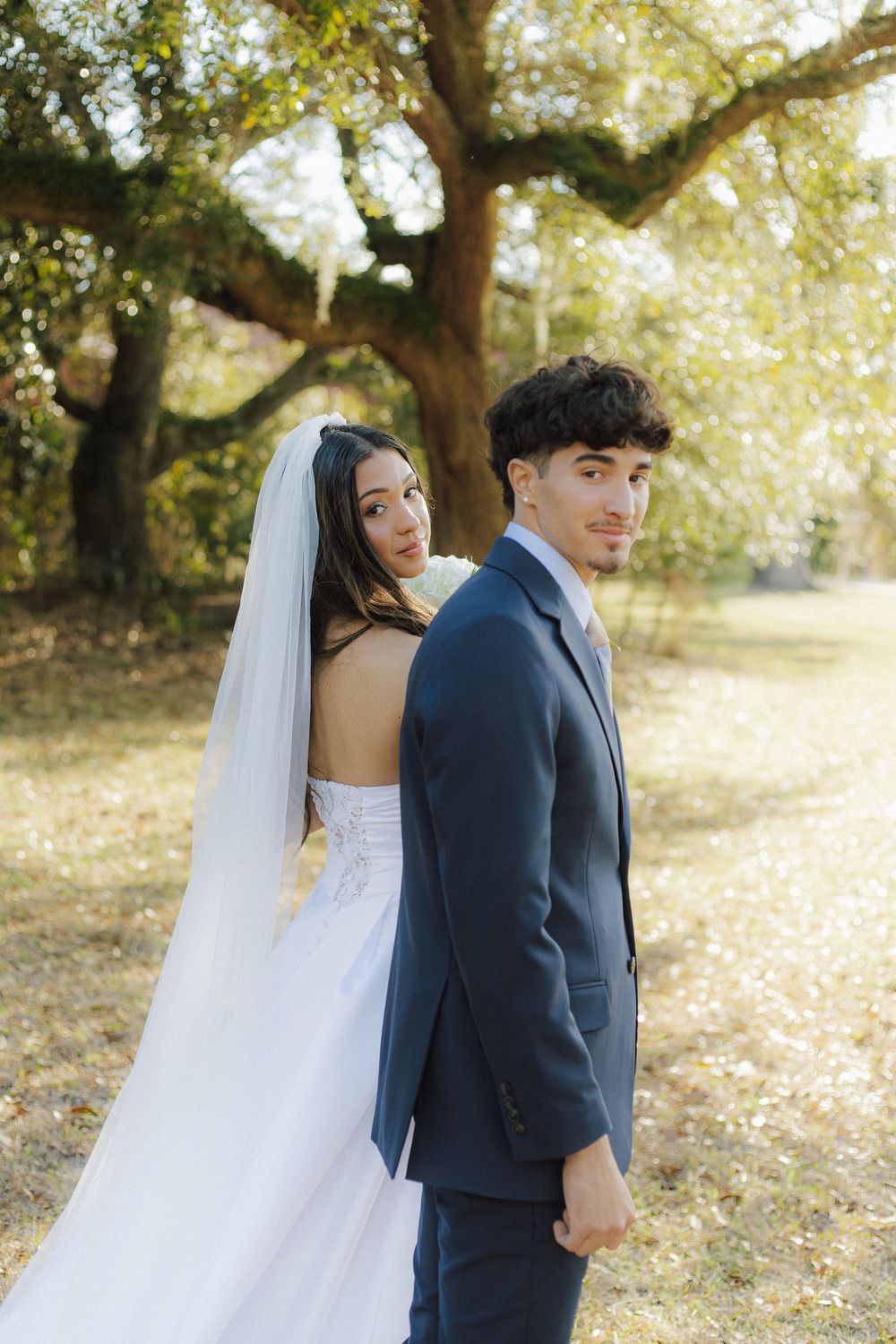A bride in white gown and groom in navy suit pose together beneath sprawling tree branches in golden sunlight.