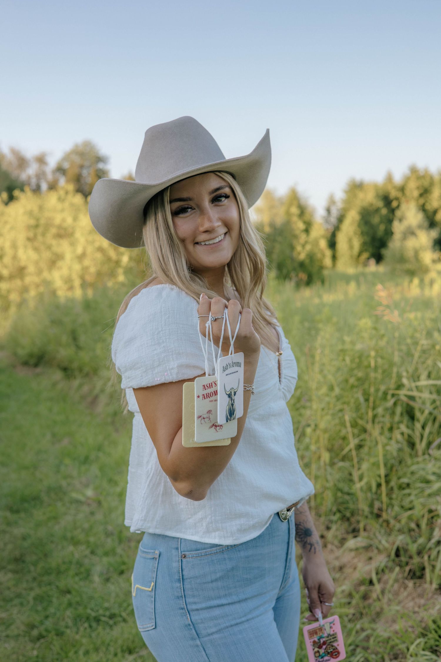 Smiling blonde woman in cowboy hat holding small product tags outdoors in green field.