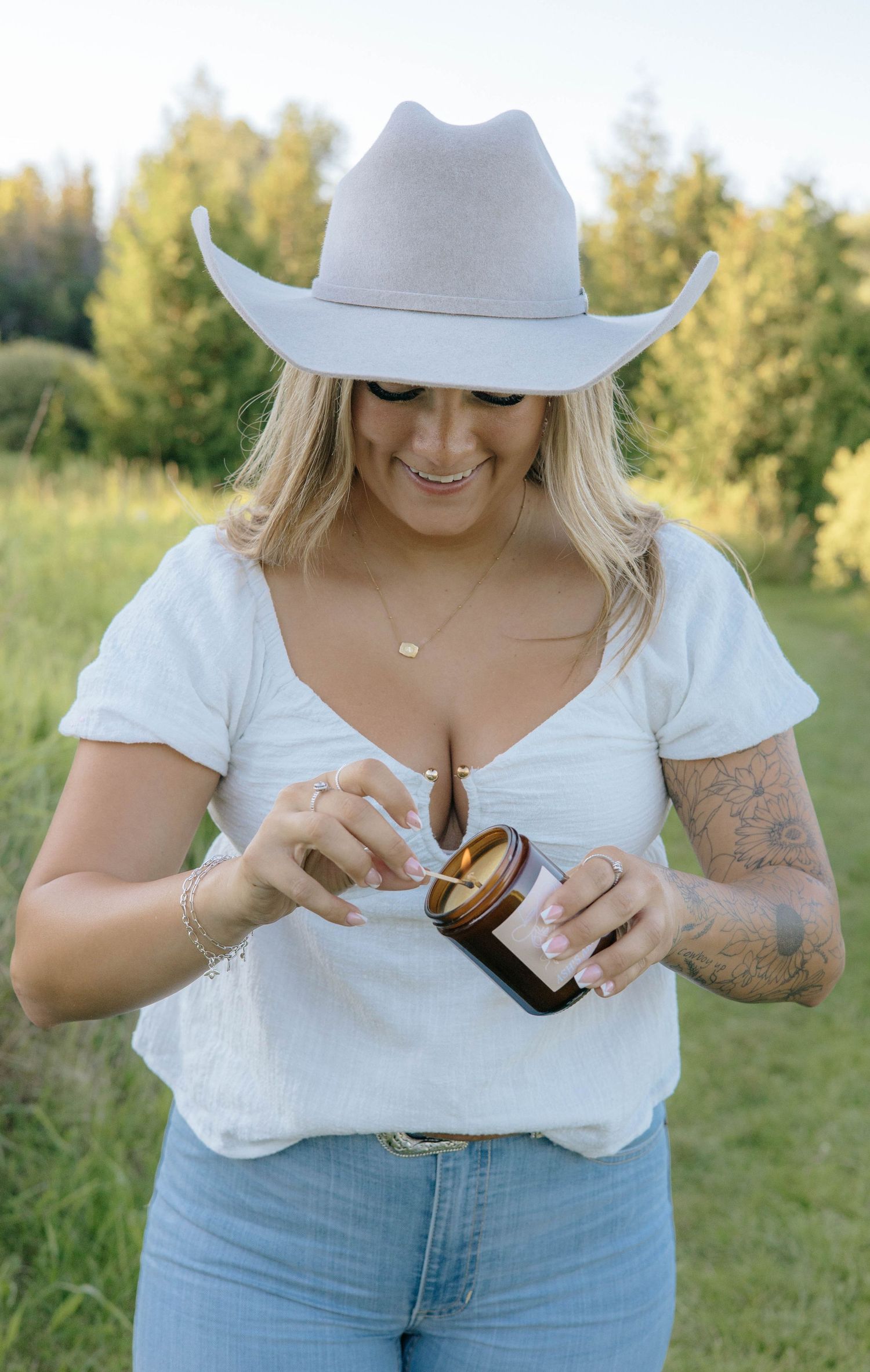Smiling blonde woman in cowboy hat trimming candle wick in amber jar outdoors.
