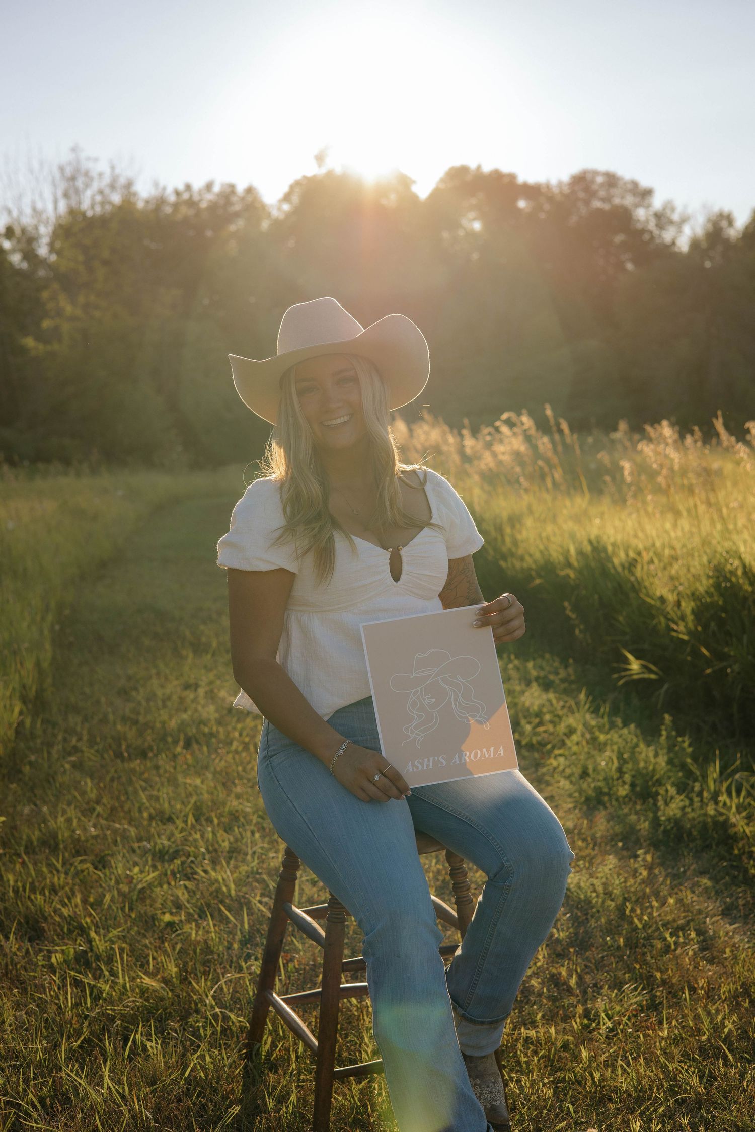 Smiling blonde woman in cowboy hat holding Ash's Aroma sign in sunny green field.