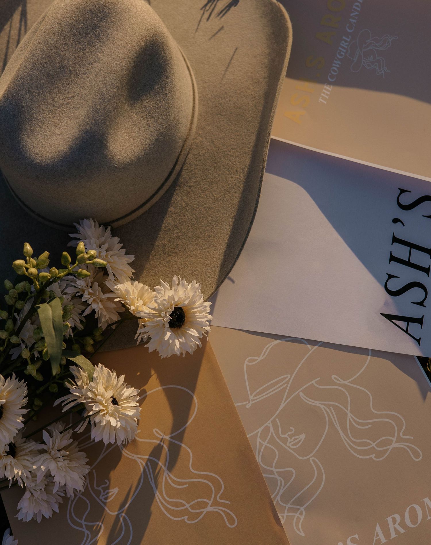 Beige cowboy hat with white flowers and Ash's Aroma branded packaging in warm light.