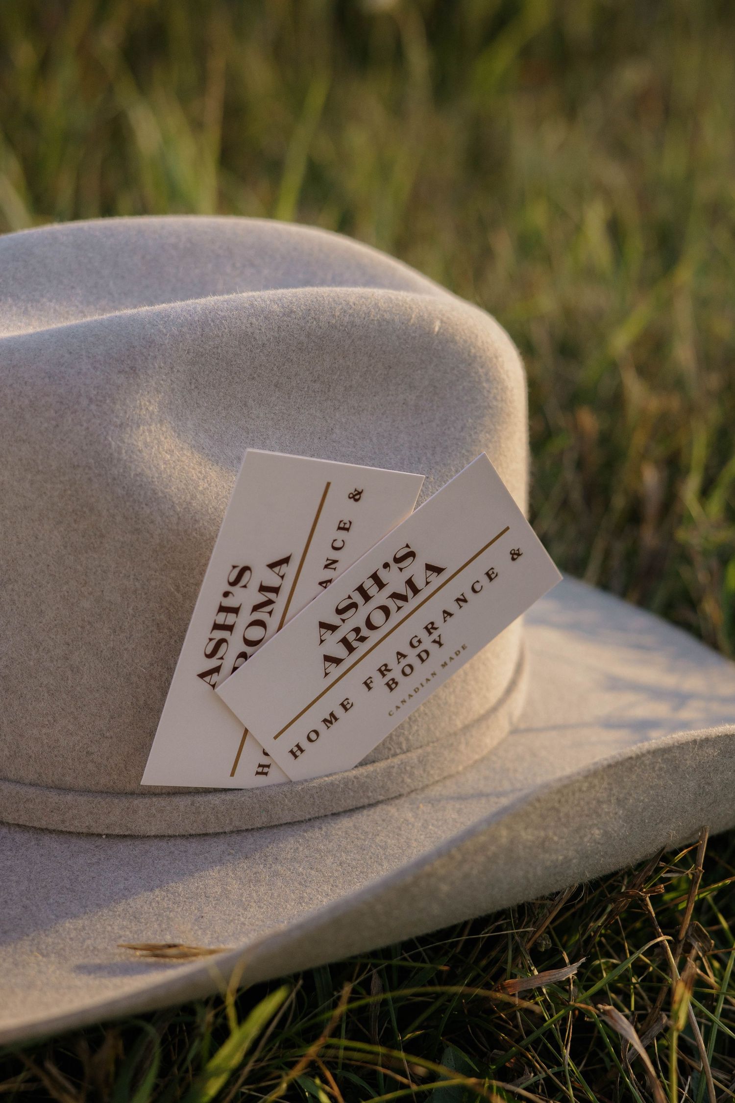 Two Ash's Aroma home fragrance business cards resting on a beige cowboy hat in grass.
