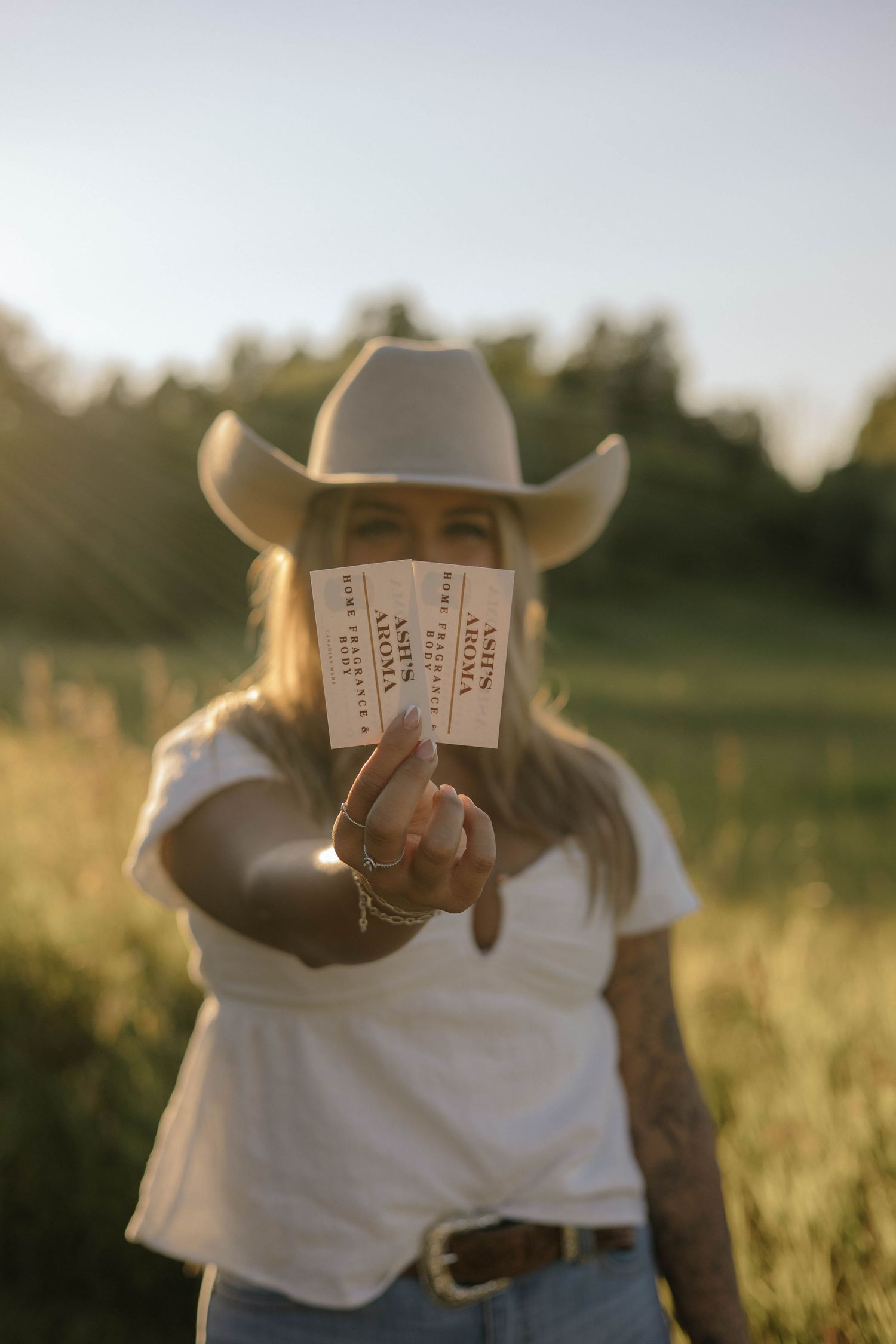 Blonde woman in white cowboy hat holding two Ash's Aroma tickets toward camera in field.