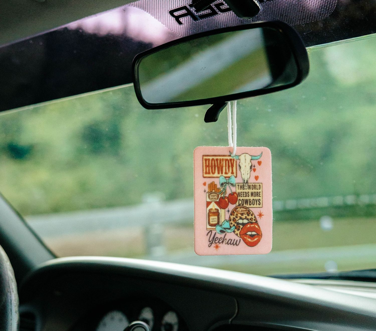 Colorful Howdy Yeehaw car air freshener hanging from rearview mirror while driving.