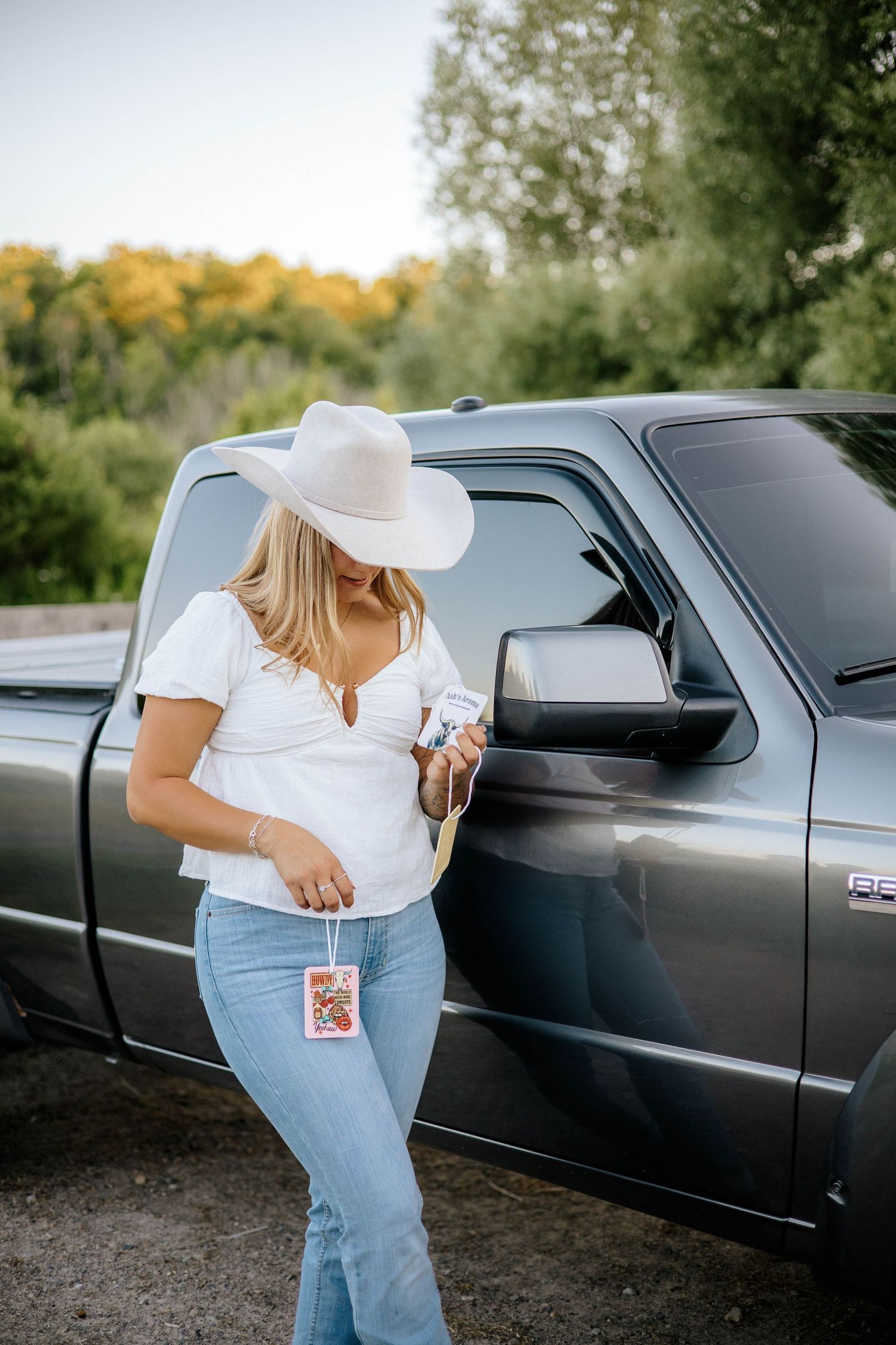 Blonde woman in white cowboy hat holding Ash's Aroma product beside gray pickup truck.