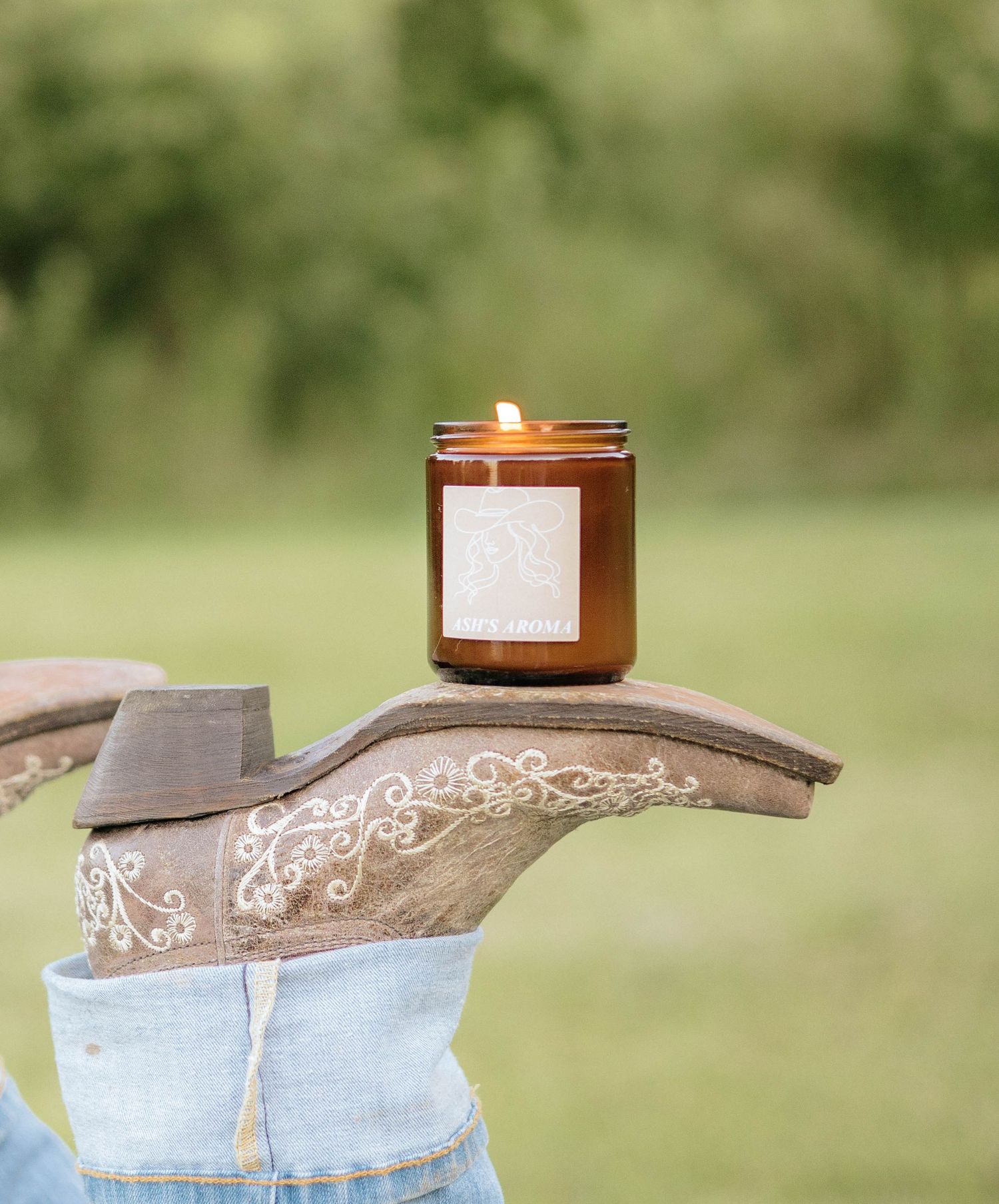 Lit amber jar candle balanced on embroidered cowboy boot toe outdoors.