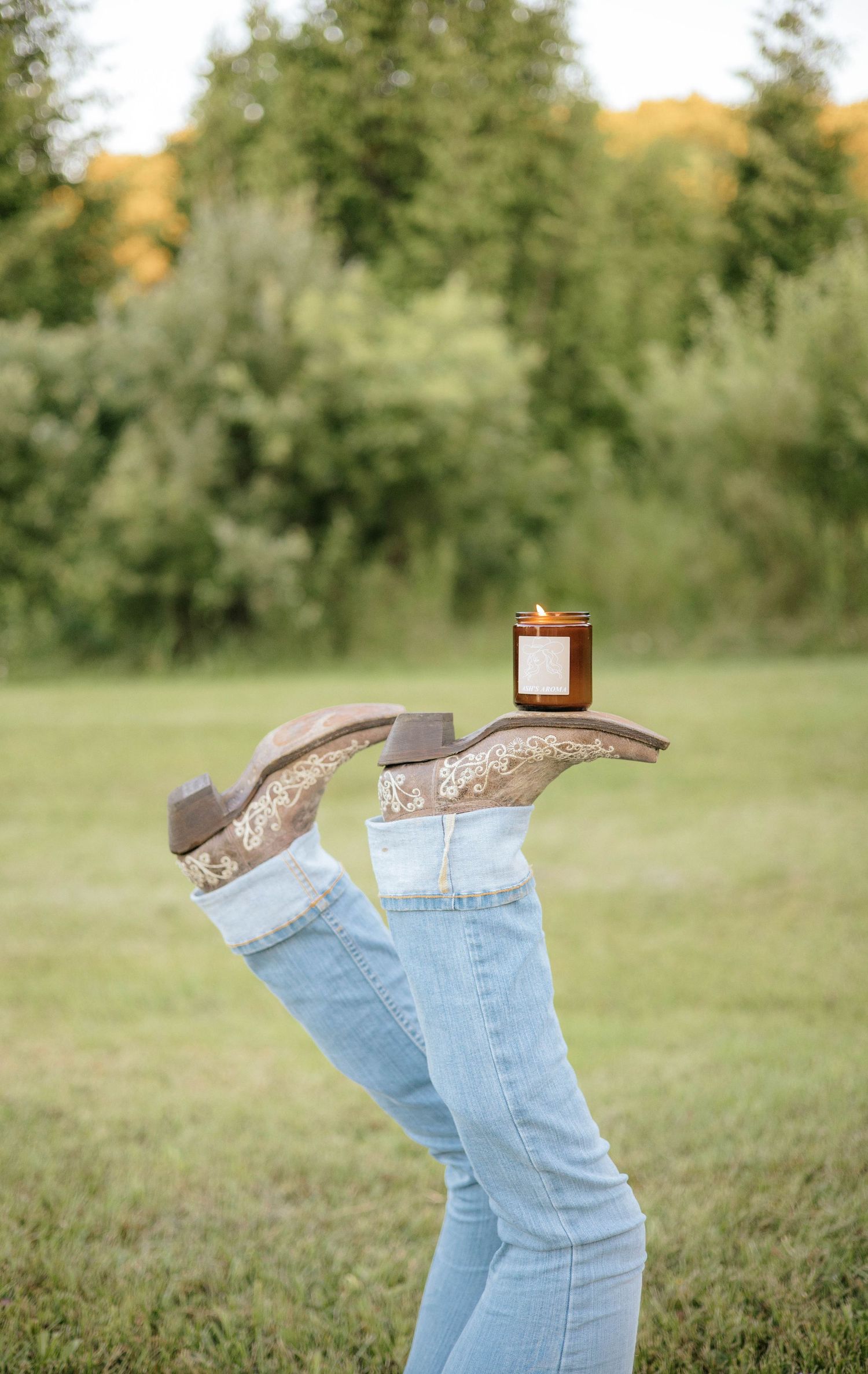 Legs raised in cowboy boots balancing a lit amber jar candle in grassy field.