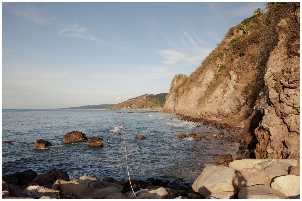 Rocky coastal cliffside with blue ocean waters and distant headland under a clear sky.