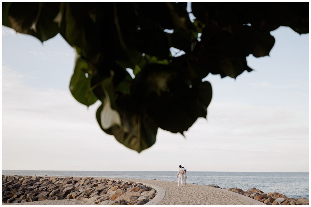 Couple walking on a rocky beach shoreline framed by tropical leaves with calm ocean horizon.