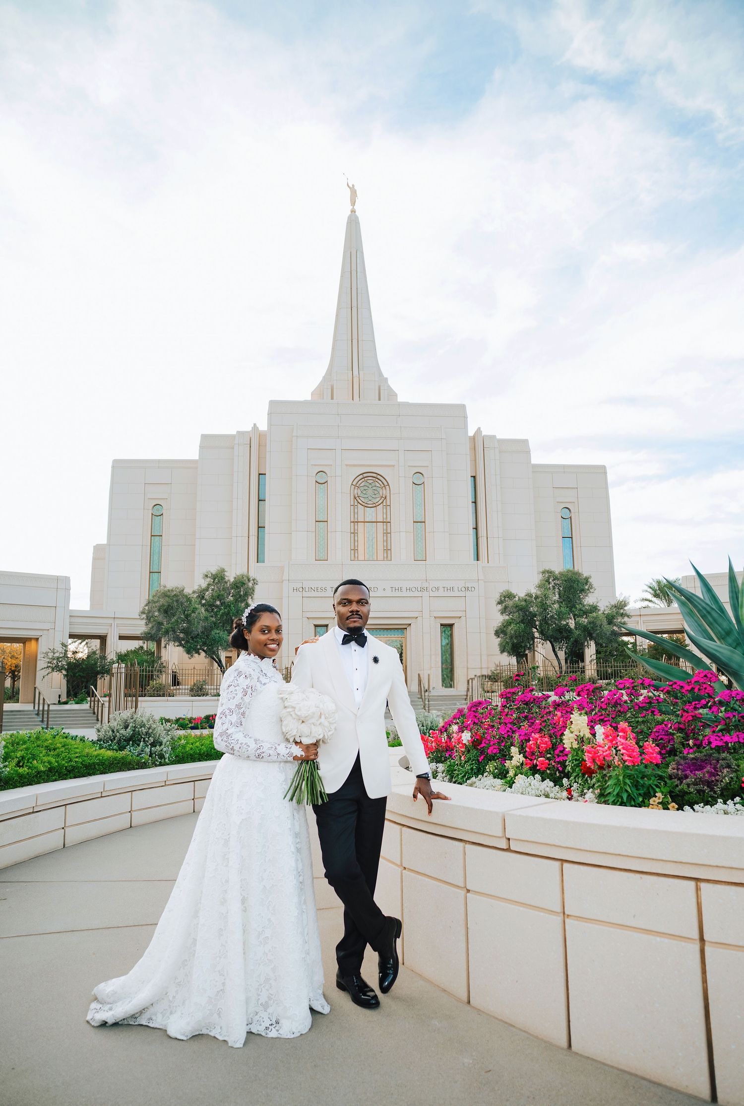 Bride and groom posing in front of a white LDS temple with a golden spire and colorful flowers.