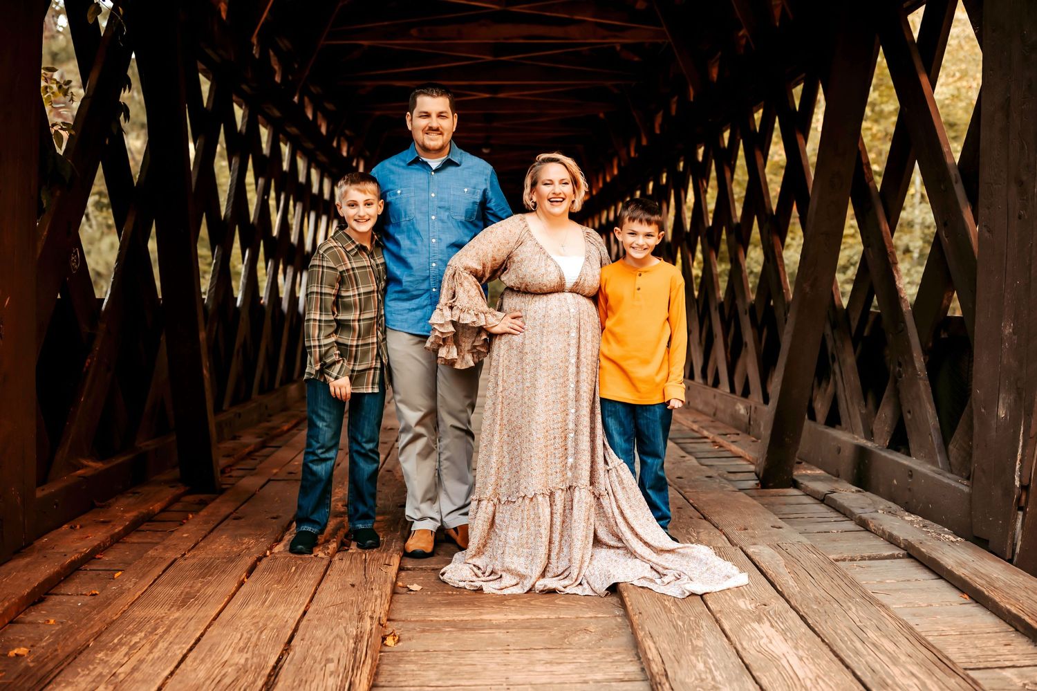 Family of four poses on wooden covered bridge, pregnant mother in floral gown with husband and two sons.
