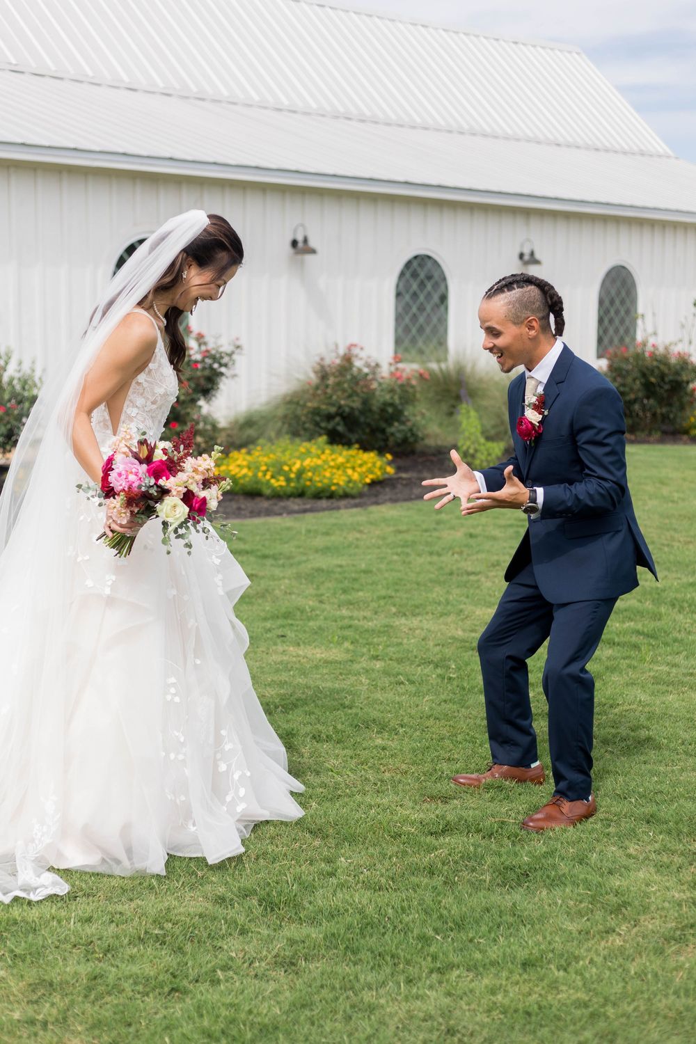 Groom in navy suit reacts with joy seeing bride in white gown holding colorful bouquet outside white barn venue.