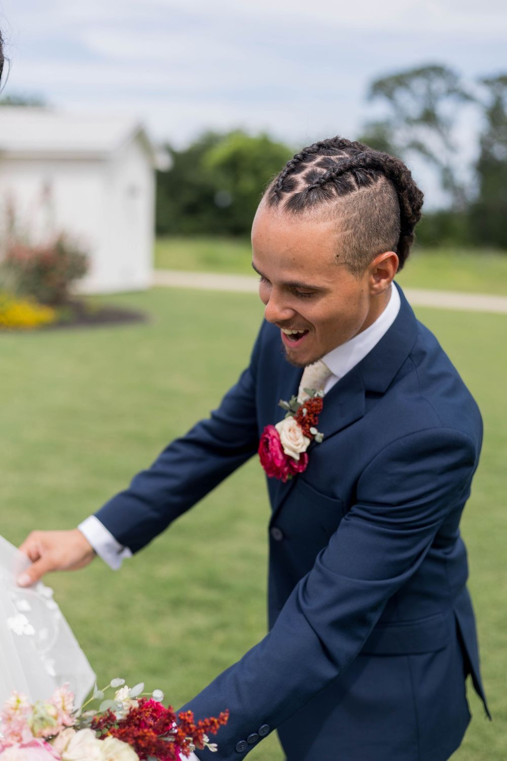Smiling groom in navy suit with red boutonniere reaching toward wedding cake outdoors.
