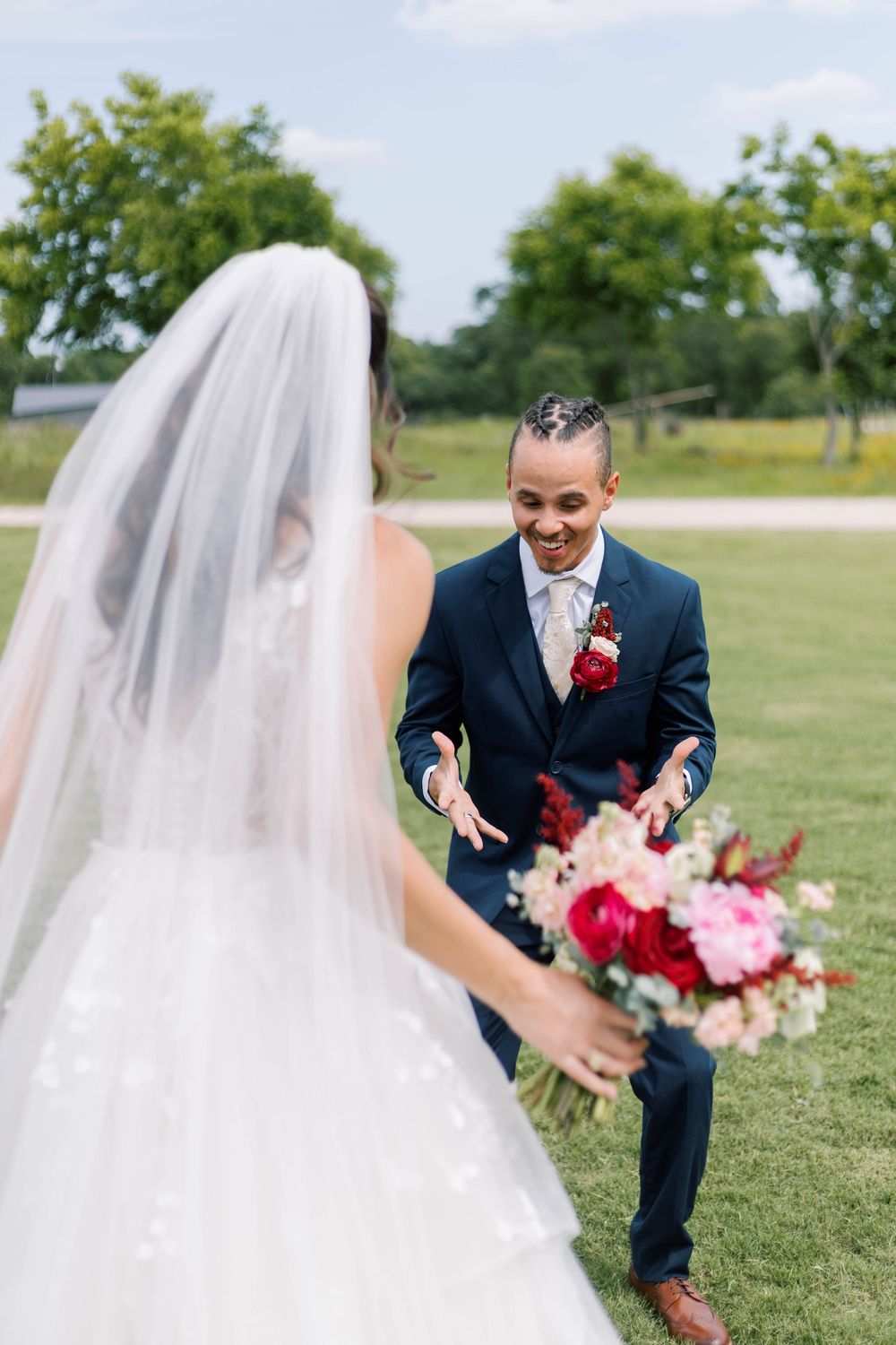 Groom in navy suit reacts with joy seeing bride in white gown and veil holding pink bouquet outdoors.