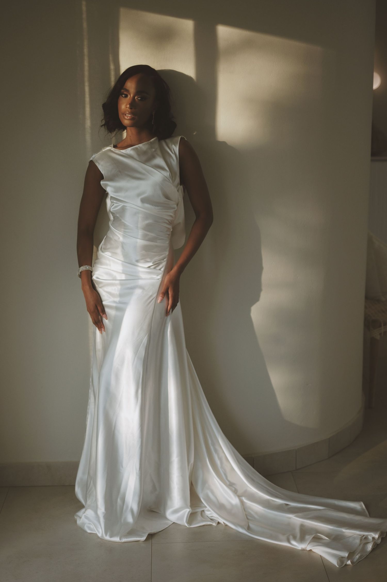 Elegant bride in sleek white satin mermaid gown poses against wall in dramatic natural light.