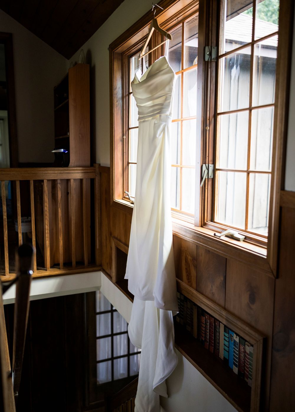 White wedding dress hanging on a hanger by a sunlit wooden window in a rustic cabin interior.