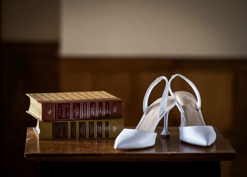 White satin bridal heels displayed beside vintage leather-bound books on a wooden table.