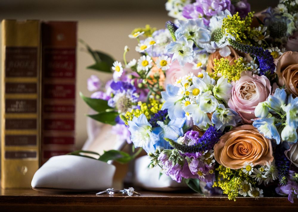 Elegant wedding bouquet with roses, hydrangeas, and daisies beside vintage books and a white vase on a wooden shelf.