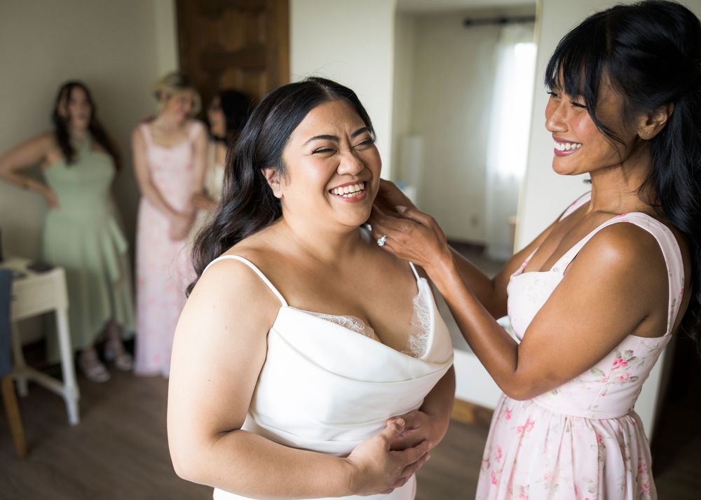 Bride smiling joyfully as bridesmaid adjusts her necklace while bridesmaids look on in background.