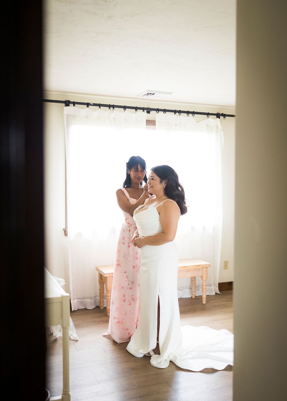 Bride in white gown gets help from bridesmaid in floral dress during wedding preparations.