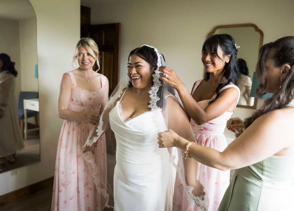 Bridesmaids help a smiling bride adjust her lace veil while getting ready in a bright room before the wedding.